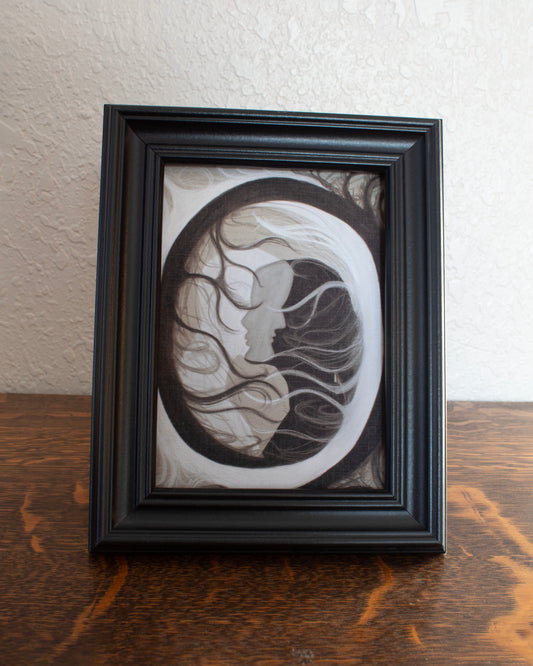 An abstract circle with black and white figures overlapping in the middle on a cream background. Framed artwork with a circular design on a wooden surface and textured wall background