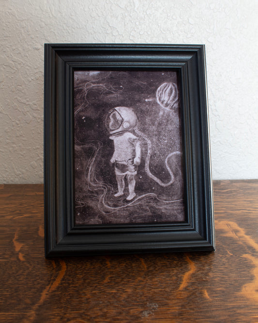 Illustration of a child astronaut in space with a dark background. Framed artwork of an astronaut on a textured wall background on a wooden table.