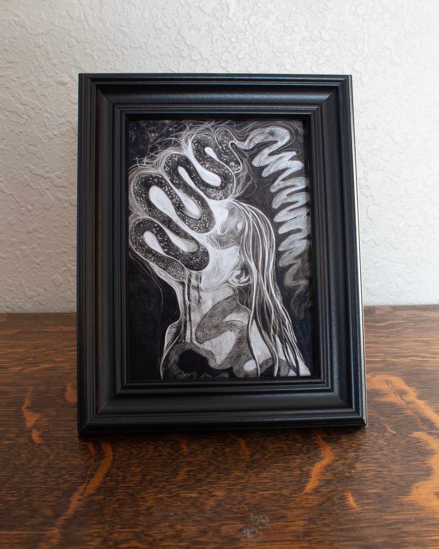 Black and white abstract artwork with flowing lines and patterns of a illuminated snake and its shedding skin. Framed artwork of a stylized face on a textured wall background on a wooden table.