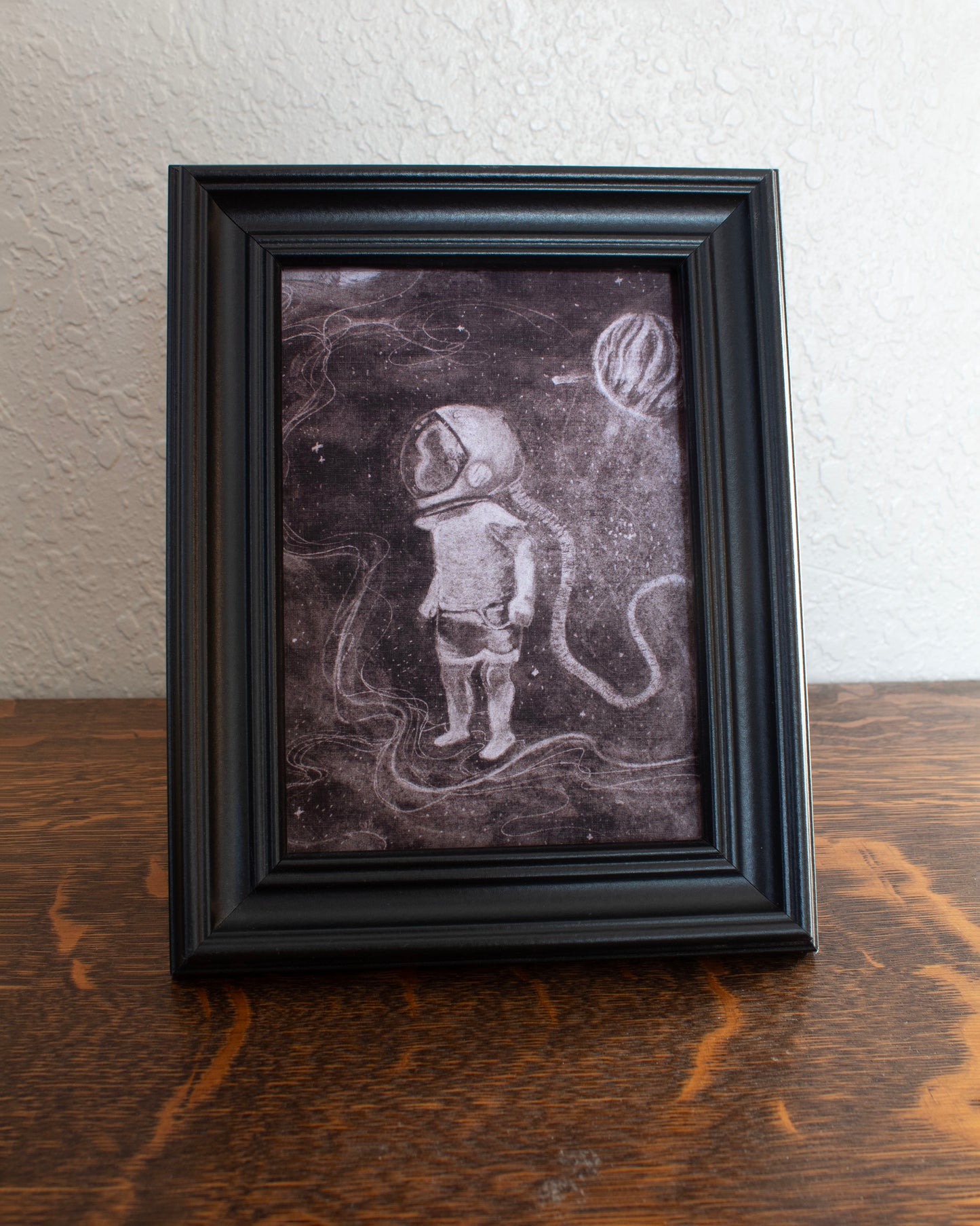 Illustration of a child astronaut in space with a dark background. Framed artwork of an astronaut on a textured wall background on a wooden table.