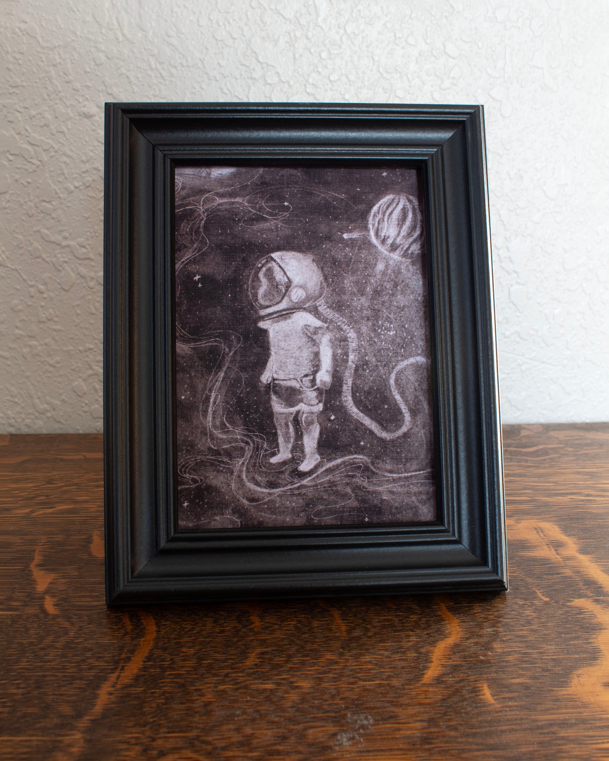 Illustration of a child astronaut in space with a dark background. Framed artwork of an astronaut on a textured wall background on a wooden table.