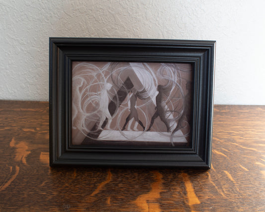 Abstract artwork with swirling lines and silhouettes on a brown background. Framed black and white artwork on a wooden surface with a white wall background