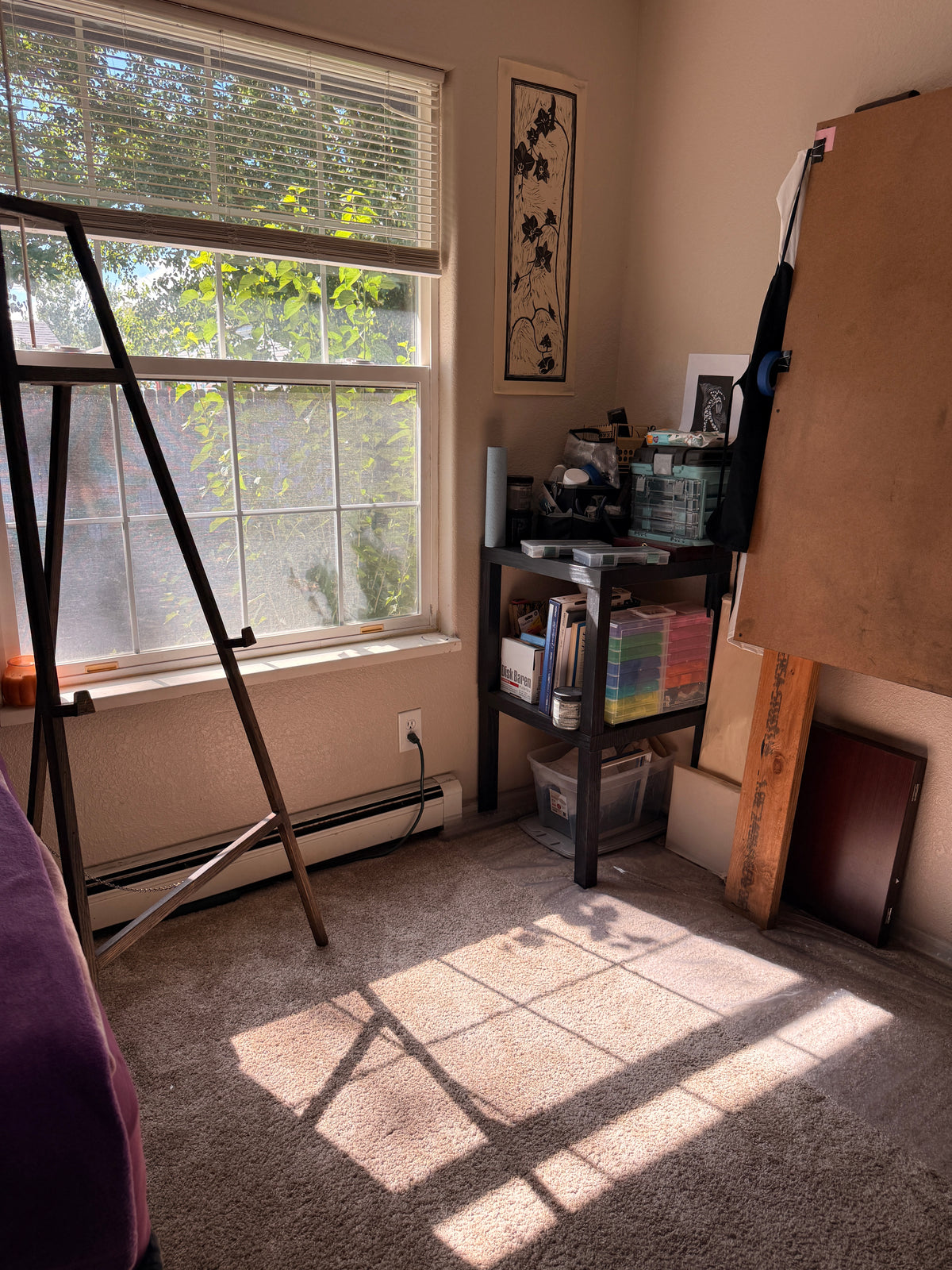 A sunlit room corner with a large window, carpeted floor, shelving, and an easel or canvas stand leaning against the wall.