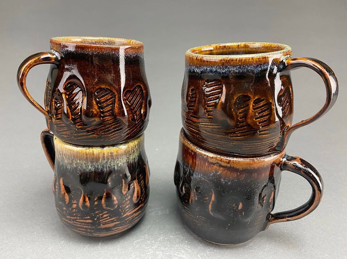 Four handmade ceramic mugs stacked in two pairs of two, featuring a glossy dark brown and black glaze with a lighter tan rim and textured relief patterns, set against a neutral gray background.