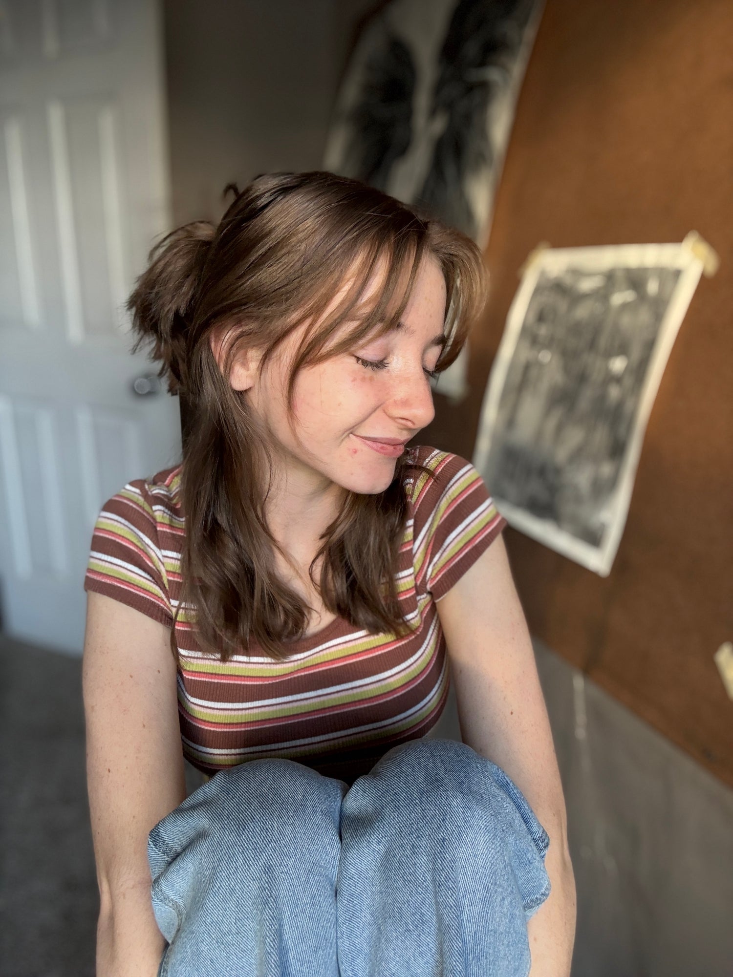 A woman with light brown hair in a half-up bun wearing a brown, red, and yellow striped t-shirt and light blue pants, sitting in an indoor setting of an art studio, with artwork behind her.