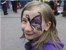 A young girl with butterfly face paint on her forehead and cheek, wearing a purple top, photographed outdoors on a paved surface with blurred people in the background.
