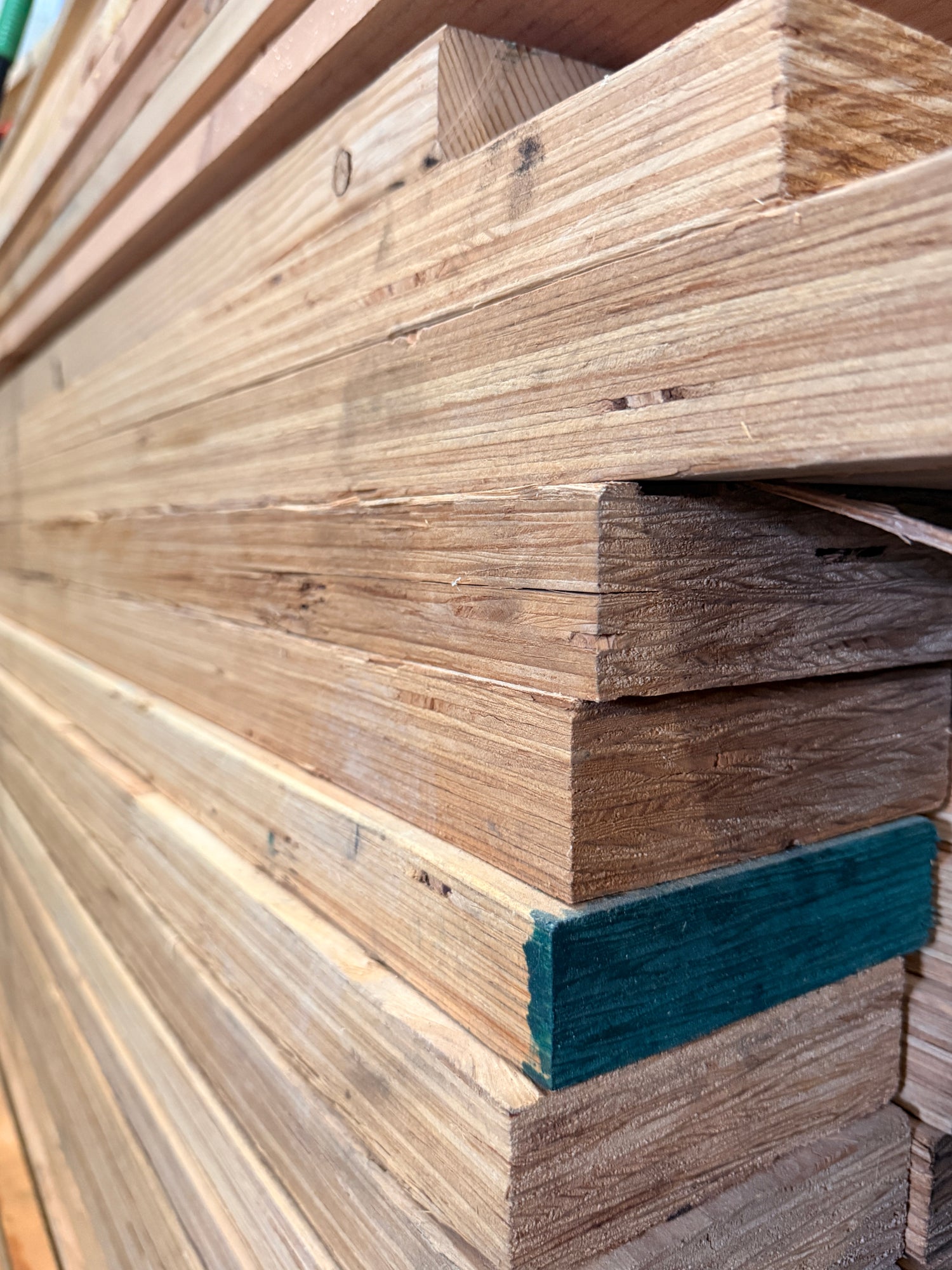Stacked natural wood planks with visible grain and a small dark teal protective coating on one board, photographed in a close-up stack view.